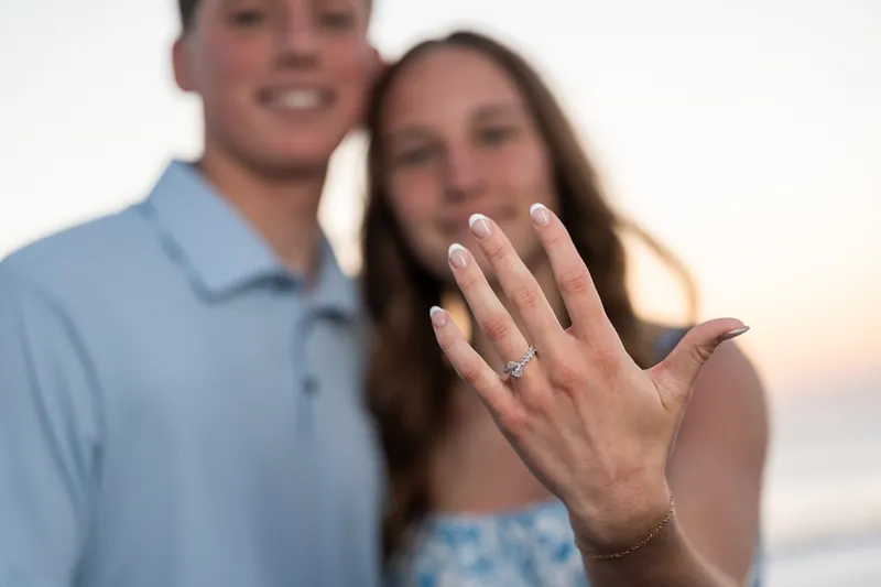 Teigan displays her engagement ring to the camera with a bright smile while Alex stands beside her on the beach at dusk.