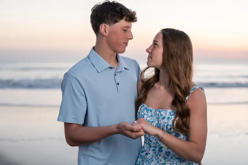 Alex and Teigan hold hands and gaze at each other on the beach during golden hour, with the ocean visible behind them.