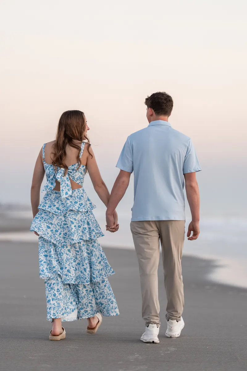 Alex and Teigan walk hand-in-hand along the wet sand of the beach at sunset with the ocean in the background.