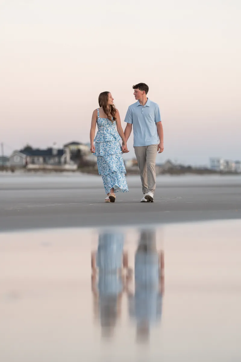 Alex and Teigan walk along the reflective wet sand on the beach at sunset, their reflections visible on the water below them.