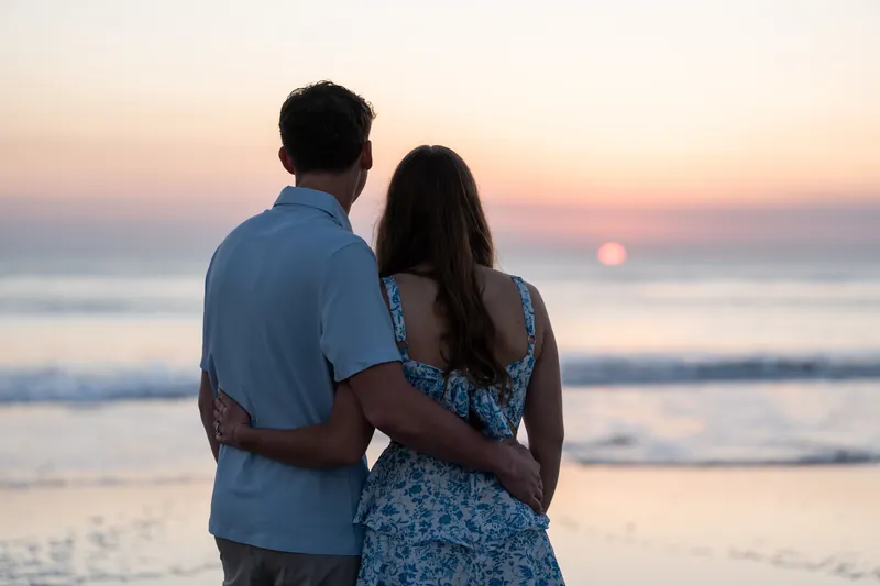 Alex and Teigan stand together facing the ocean at sunset, with Alex wearing a light blue shirt and Teigan in a blue floral dress as the sun descends over the water behind them.