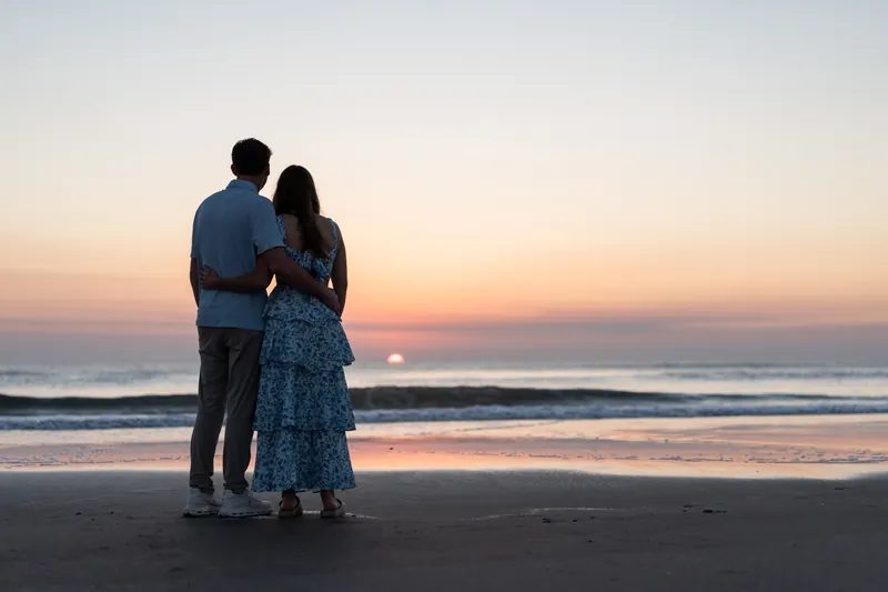 Alex and Teigan embrace while standing on the beach during golden hour, with soft waves and a pink-orange sky reflected on the wet sand.
