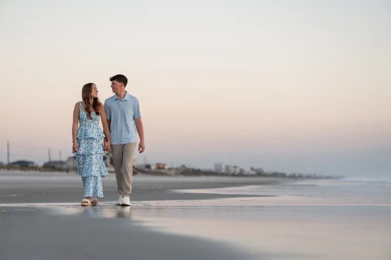 Alex and Teigan walk hand in hand along the shoreline at dusk, with Teigan in a blue floral dress and Alex in a gray shirt as the sky glows with sunset colors.
