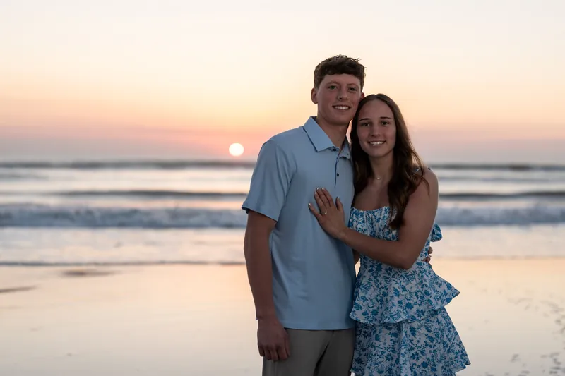 Alex wraps his arm around Teigan's shoulders as they stand on the beach at sunset, both smiling with the sun low on the horizon behind them.