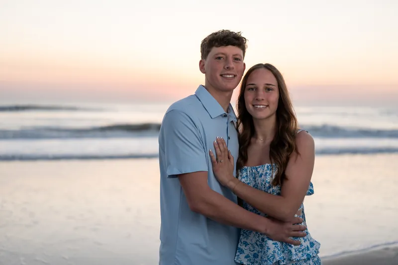 Alex and Teigan pose together on the beach with his arm around her waist, both looking at the camera with the ocean and pink sunset sky behind them.