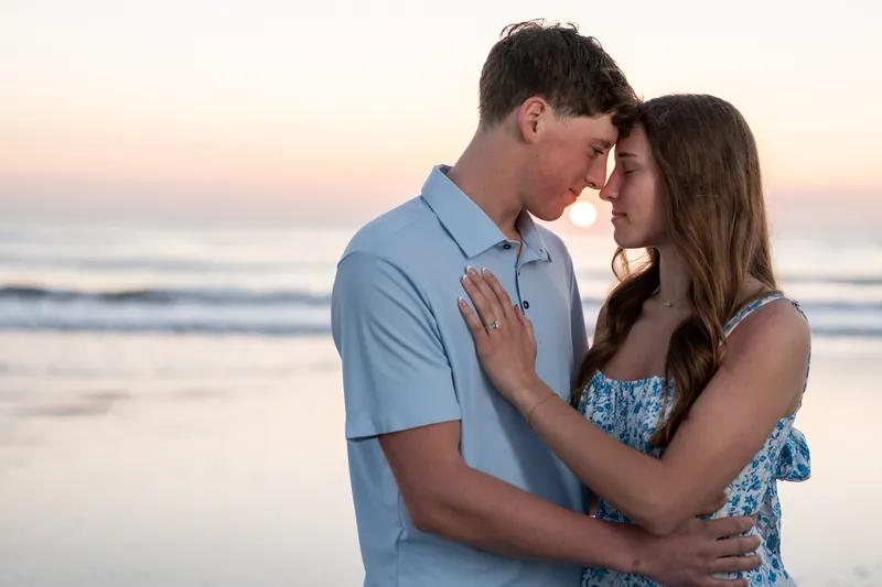 Alex and Teigan face each other close together on the beach, with their foreheads nearly touching and the golden sunset reflecting off the water in the background.