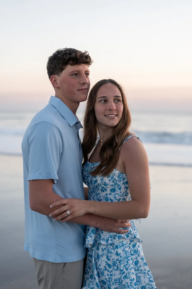 Alex and Teigan stand together on the beach at dusk, with Alex's arm around Teigan's waist.