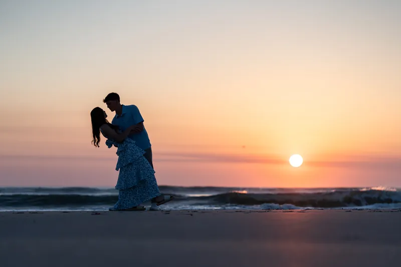 Alex dips Teigan during a kiss as the sun sets over the ocean behind them.