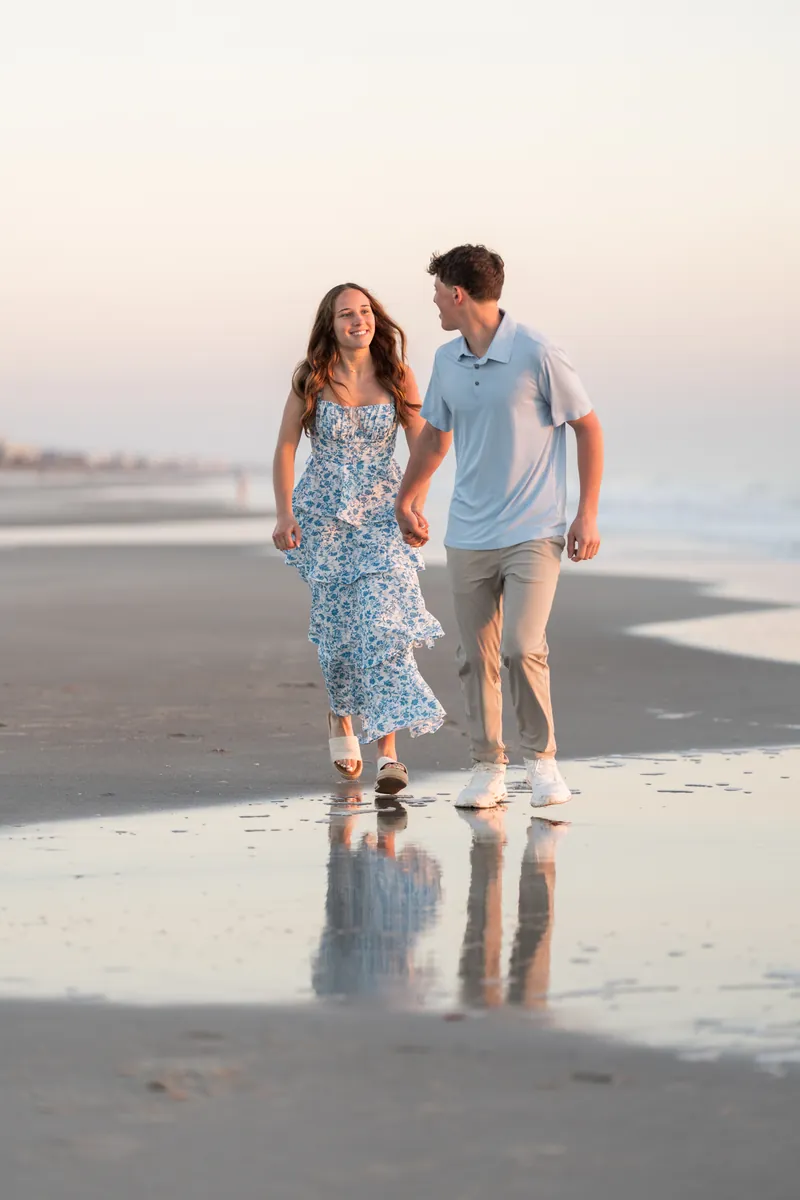 Alex and Teigan walk hand in hand along the wet sand at the shoreline during golden hour.