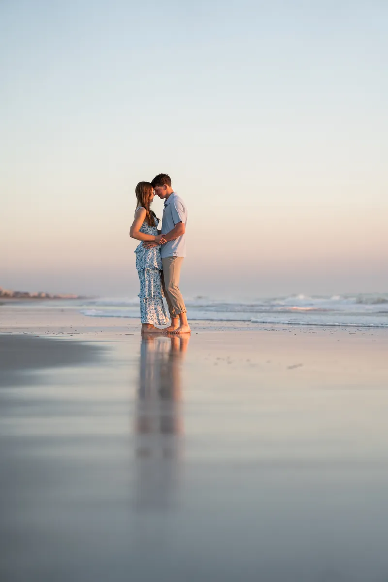 Alex lifts Teigan in an embrace while they stand in shallow water on the beach at sunset.