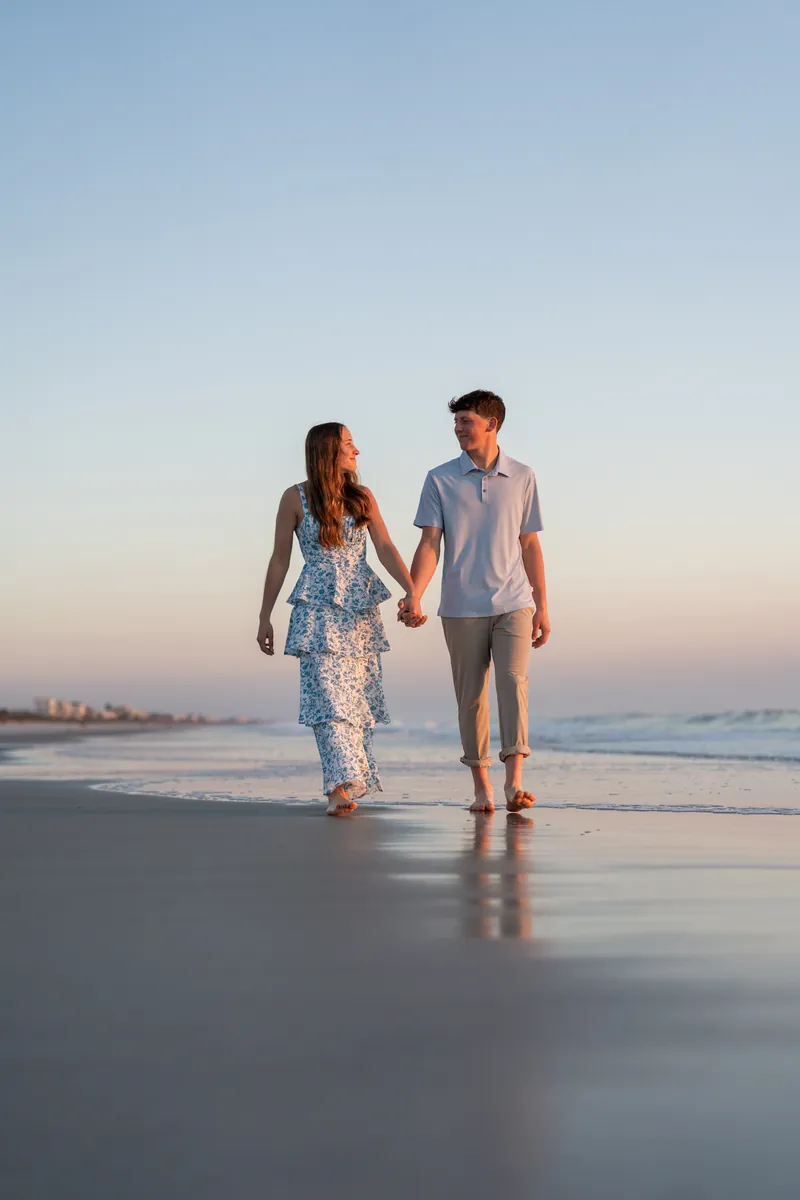 Alex and Teigan walk barefoot along the wet sand holding hands as the sun sets over the water.