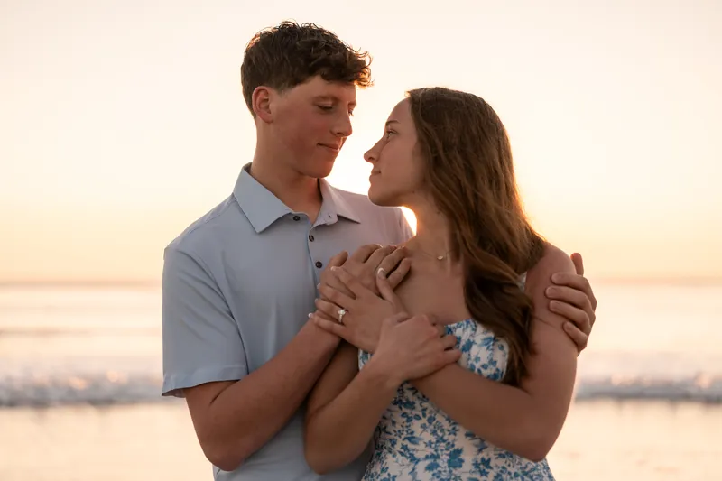 Alex holds Teigan's hands at chest level while facing her against a sunset ocean backdrop.
