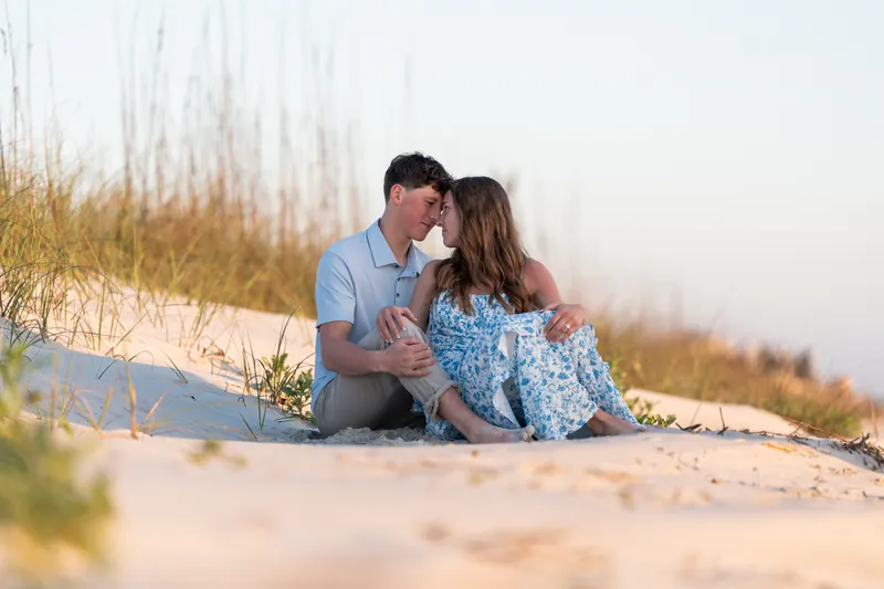 Alex and Teigan sit together on sand dunes surrounded by beach grass during golden hour.