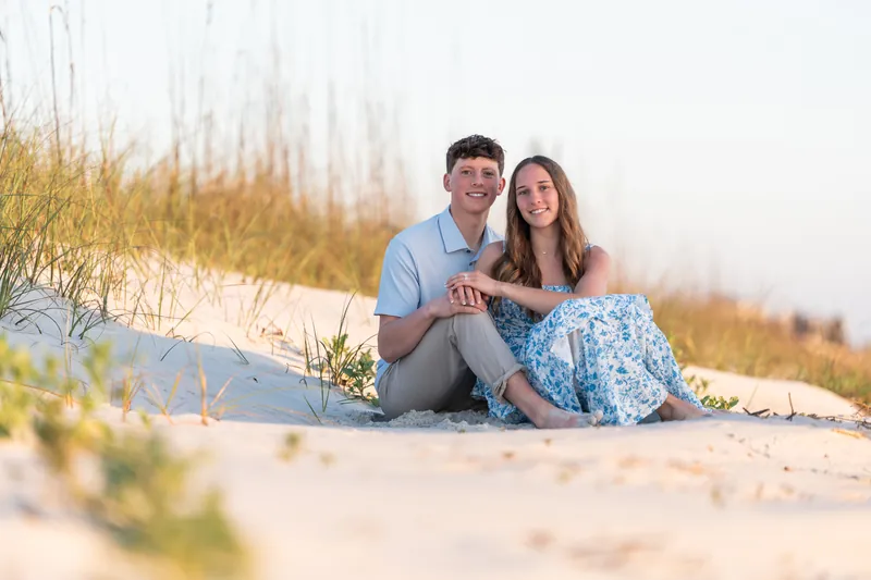 Alex and Teigan sit close on the sand with dune grass and water visible behind them at dusk.