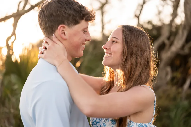 Alex and Teigan face each other smiling with their arms around each other's necks under a twisted tree at sunset.