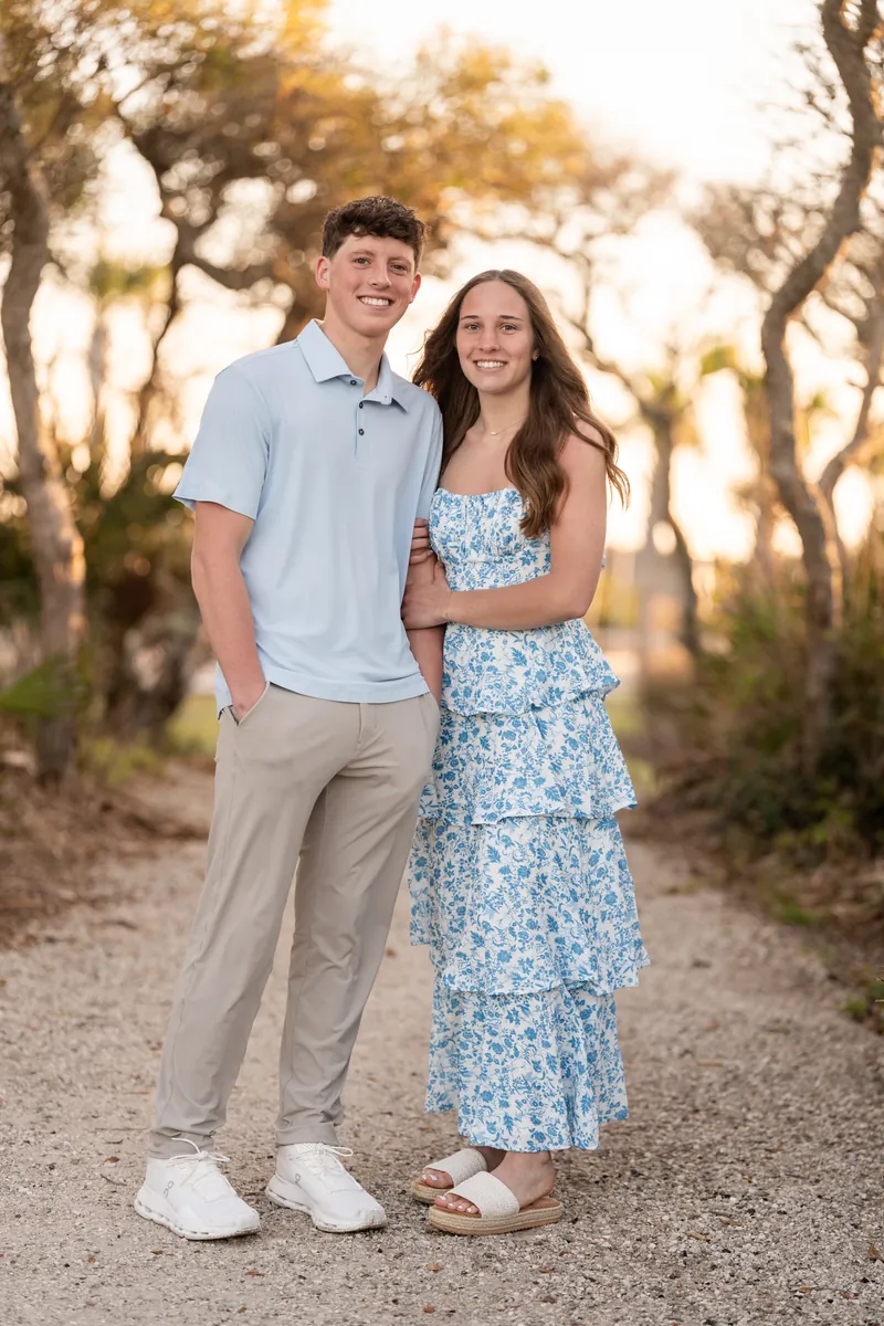 Alex and Teigan stand together on a tree-lined dirt path, both smiling at the camera during golden hour.