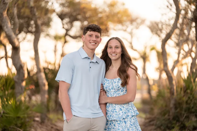 Alex and Teigan embrace while standing on a shaded dirt trail surrounded by tall oak trees at sunset.
