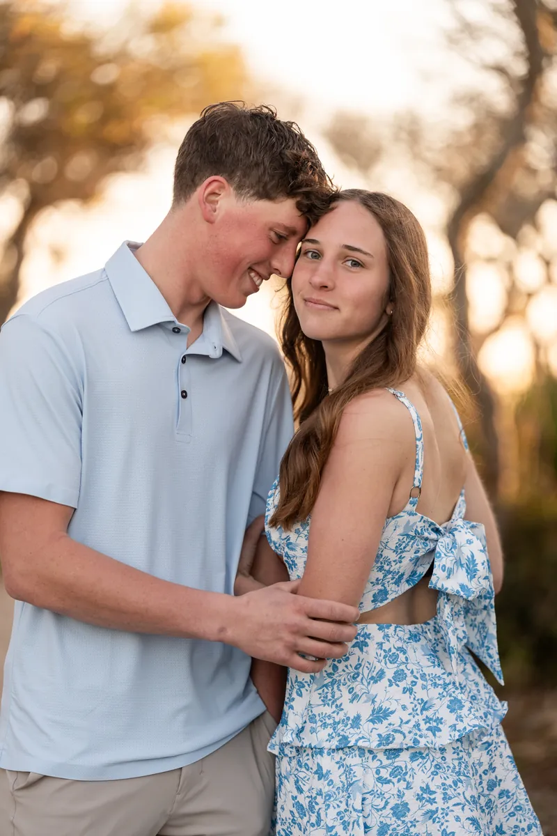 Alex rests his head against Teigan's as they stand together on a tree-lined trail during golden hour.