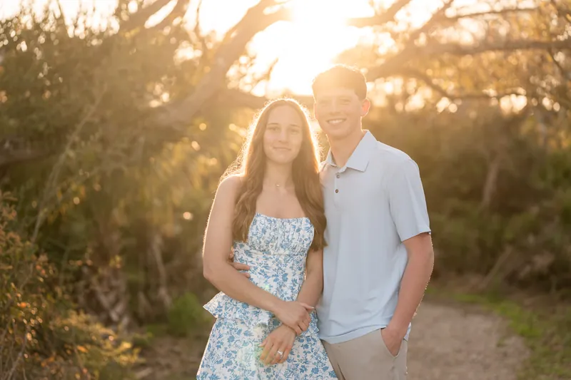 Alex and Teigan stand backlit by bright sunlight filtering through oak trees, with Teigan leaning against Alex's side.