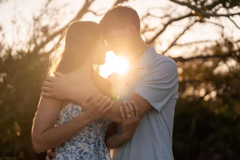 Alex and Teigan kiss backlit by golden sunlight among bare trees at sunset.
