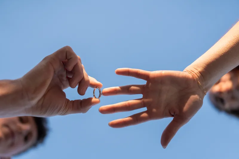 Alex slips an engagement ring onto Teigan's finger with blue sky behind them.