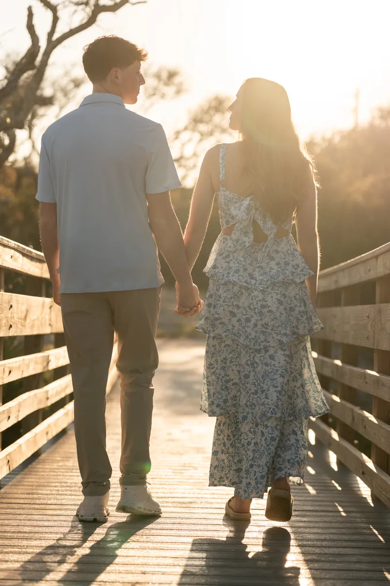 Alex and Teigan walk hand-in-hand down a wooden boardwalk at golden hour.