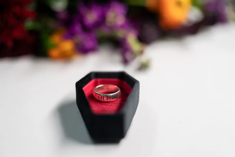 Gold wedding band with red gemstone nestled in black hexagonal box, colorful flowers blurred in background.