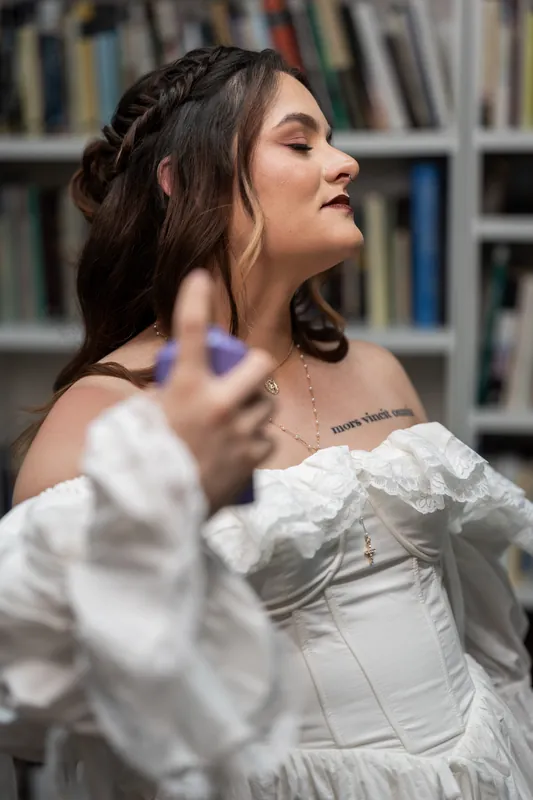 Alexandra in white off-shoulder wedding dress with braided updo, gazing upward indoors against bookshelf backdrop.