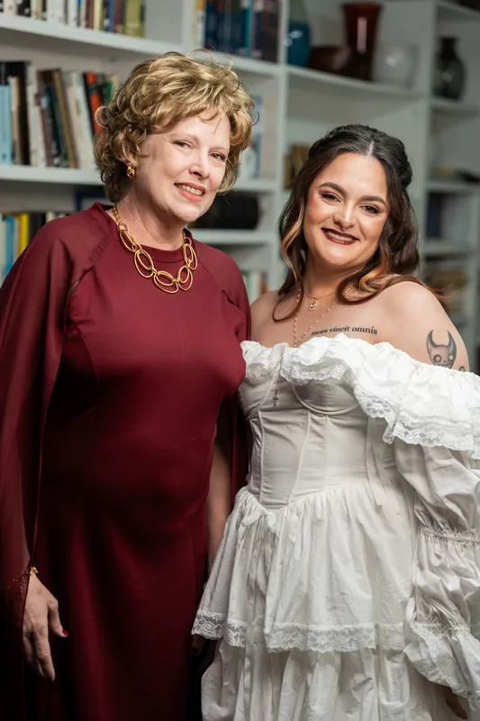 Alexandra in layered white off-shoulder gown smiles alongside woman in burgundy dress in front of home library.