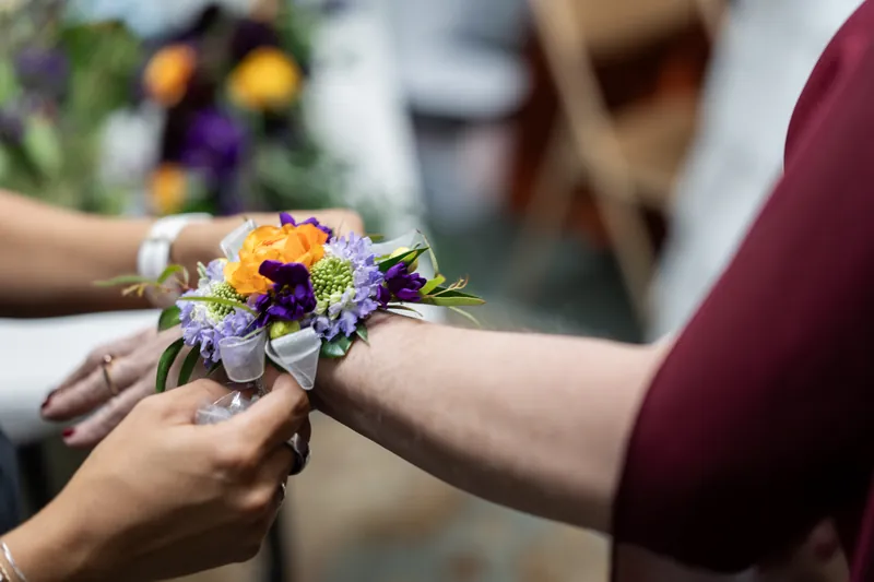 Hands exchanging yellow and purple wildflower bouquet at outdoor ceremony with burgundy-clothed guest visible.