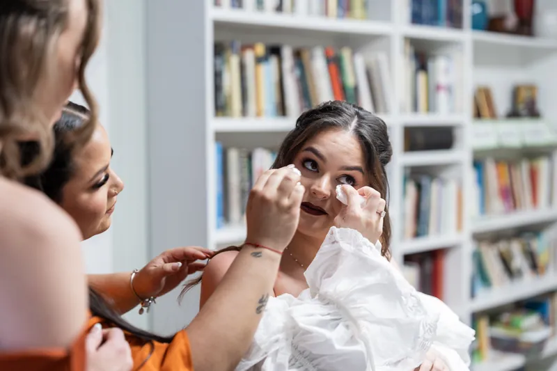 A woman in an orange shirt adjusts the bride's white veil while standing in front of built-in bookshelves.