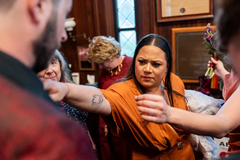 Jason reaches toward Alexandra as she gestures animatedly in an orange top during an indoor celebration.