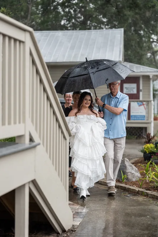 Alexandra and Jason stand under a shared black umbrella in the rain near a residential structure.