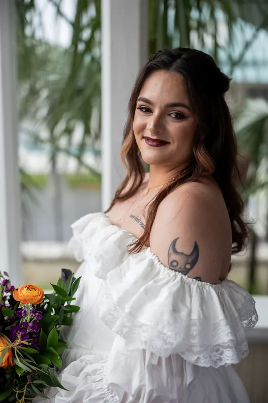 Alexandra poses in her white off-shoulder wedding dress, showing her shoulder tattoo near flowering stems.
