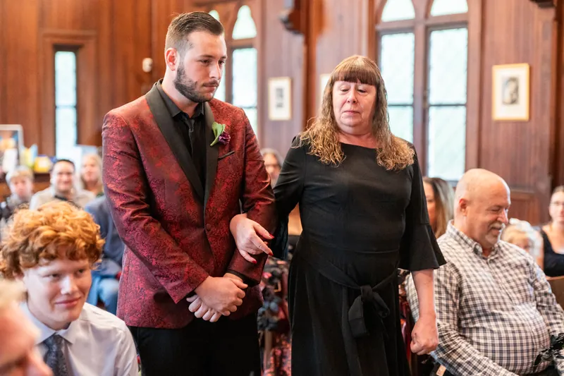 Jason in a burgundy blazer walks with a woman in black through a wood-paneled room with guests seated.