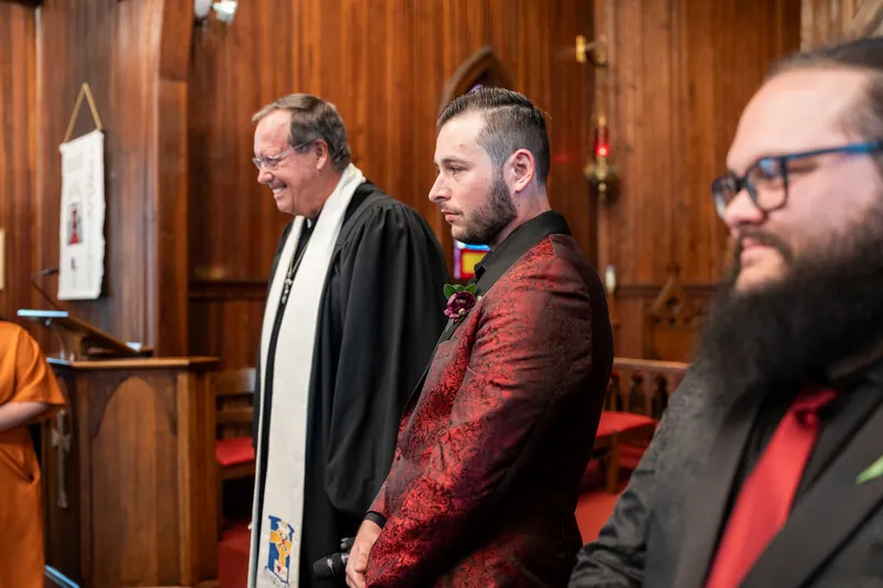 Jason stands in his burgundy jacket between two men in formal wear inside the wood-lined chapel.