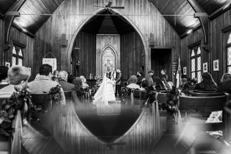 Alexandra and Jason stand before an arched doorway at Riverhouse Events as guests seated on either side watch their ceremony in black and white.