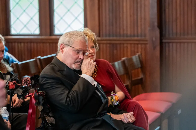 A guest in glasses and dark suit wipes his eyes during the ceremony at Riverhouse Events, visibly moved by the moment.
