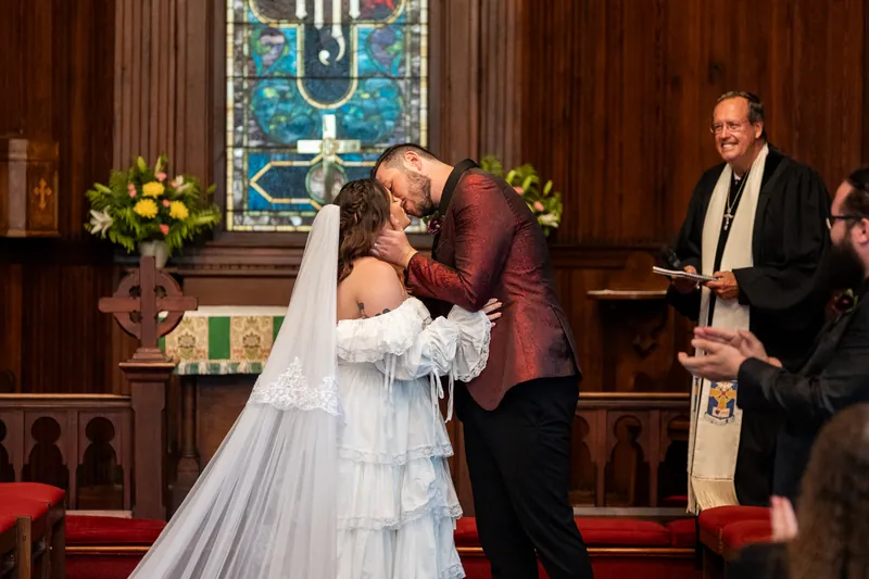 Jason kisses Alexandra on the lips as she holds her bouquet, both dressed formally in front of a stained glass window.