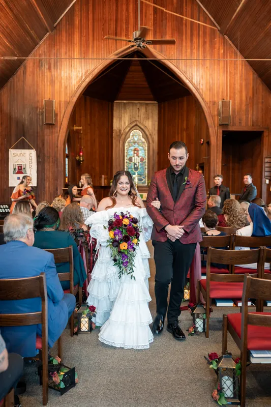Alexandra in a layered white gown stands beside Jason in a burgundy jacket while guests seated in wooden chairs look on during their ceremony.