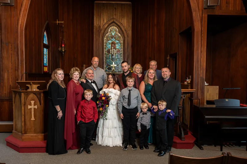Alexandra in her wedding dress poses with a large family group including children and adults dressed in formal attire inside a wooden chapel.