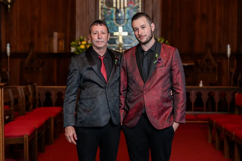 Two men stand side by side smiling at the camera on the chapel's red carpet, both wearing burgundy and black blazers.