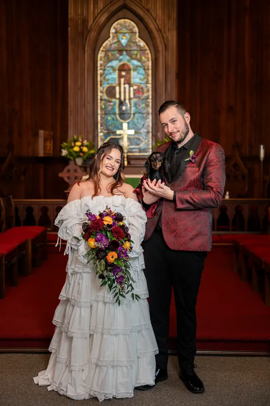 Alexandra and Jason stand before a stained-glass window in a wooden chapel, with Alexandra holding a vibrant red and purple bouquet in her tiered white gown and Jason in a burgundy velvet jacket.