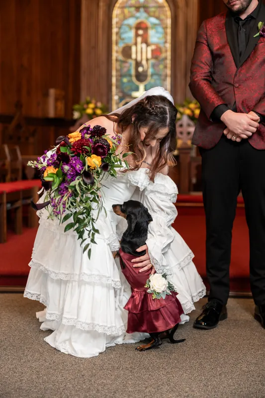 A flower girl in a white dress hugs Alexandra's bouquet while Jason stands nearby in the chapel, with the ornate stained-glass window framed behind them.