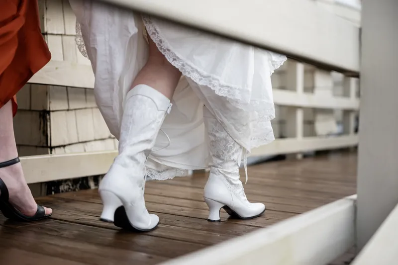 White lace-up boots peek from beneath a layered white wedding dress on a wooden deck as a guest in pink stands nearby.