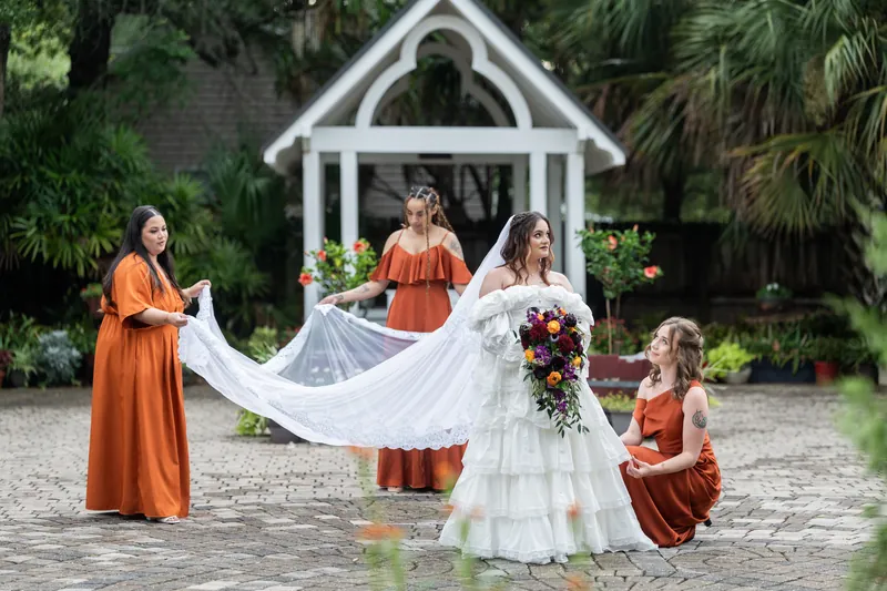 At Riverhouse Events, attendants in orange dresses hold Alexandra's flowing veil behind her as she walks on the courtyard path near a white chapel structure.
