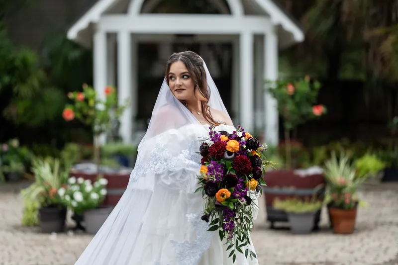 Alexandra stands alone at Riverhouse Events holding a cascading dark and gold bouquet, her long veil draped across her shoulders before the white chapel.
