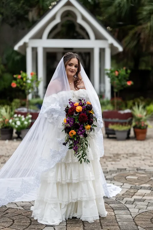 Alexandra poses on the paved courtyard at Riverhouse Events in her tiered white gown and veil, clutching her trailing floral arrangement near flowering plants and the chapel.