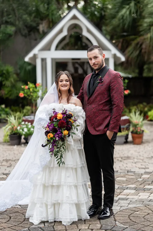 Alexandra and Jason stand at Riverhouse Events before a white chapel with tiered gardens, Alexandra in a layered white gown holding a vibrant cascade bouquet, Jason in burgundy.