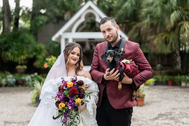 Jason holds their dog while posing alongside Alexandra at Riverhouse Events, both smiling with the white chapel visible behind them.